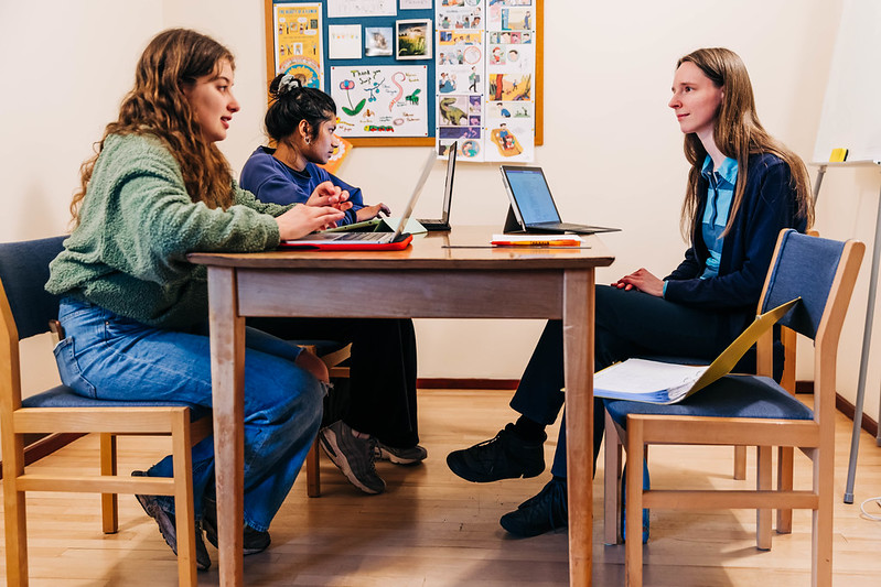 A tutor sits at a table in discussion two students. They all have laptops open.