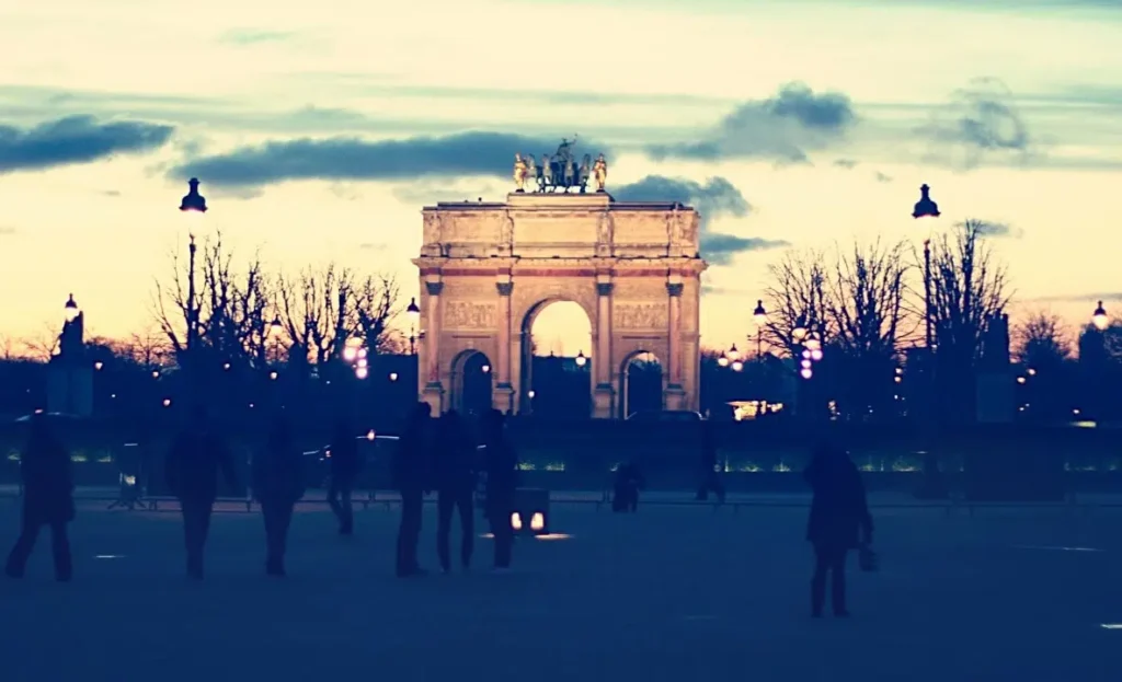 Silhouetted people walk near the Arc de Triomphe du Carrousel at dusk. The sky is a gradient of light blue to orange, with scattered clouds. Streetlights are illuminated, casting a soft glow around the historic arch.