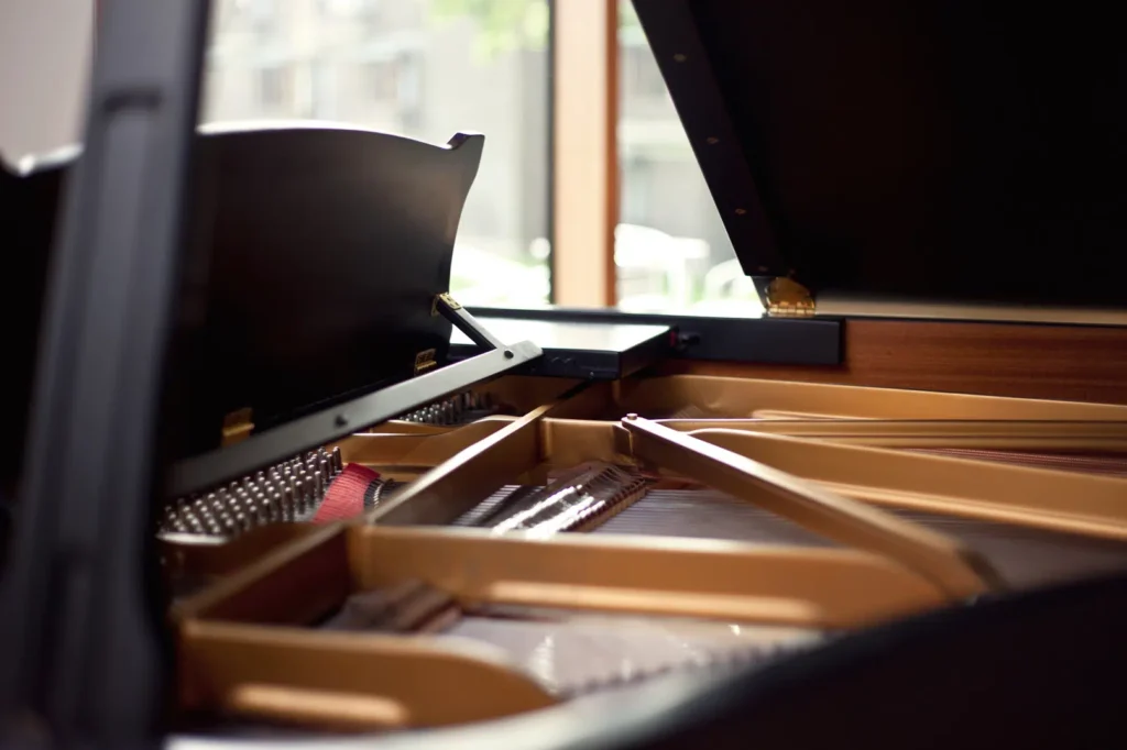 Close-up of the interior of a grand piano, showing strings, hammers, and the wooden frame. The lid is partially open, with sunlight streaming in from a window, illuminating the shiny metal components and creating a warm ambiance.