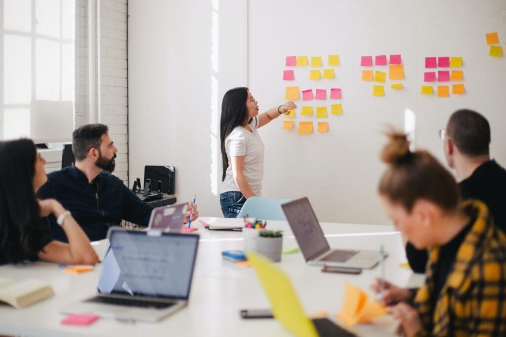 A woman stands at a whiteboard covered with colorful sticky notes, pointing at them as she speaks to four colleagues seated around a table in the sunlit office. Engaged in a design meeting, they all have laptops and notebooks ready to capture the creative flow.