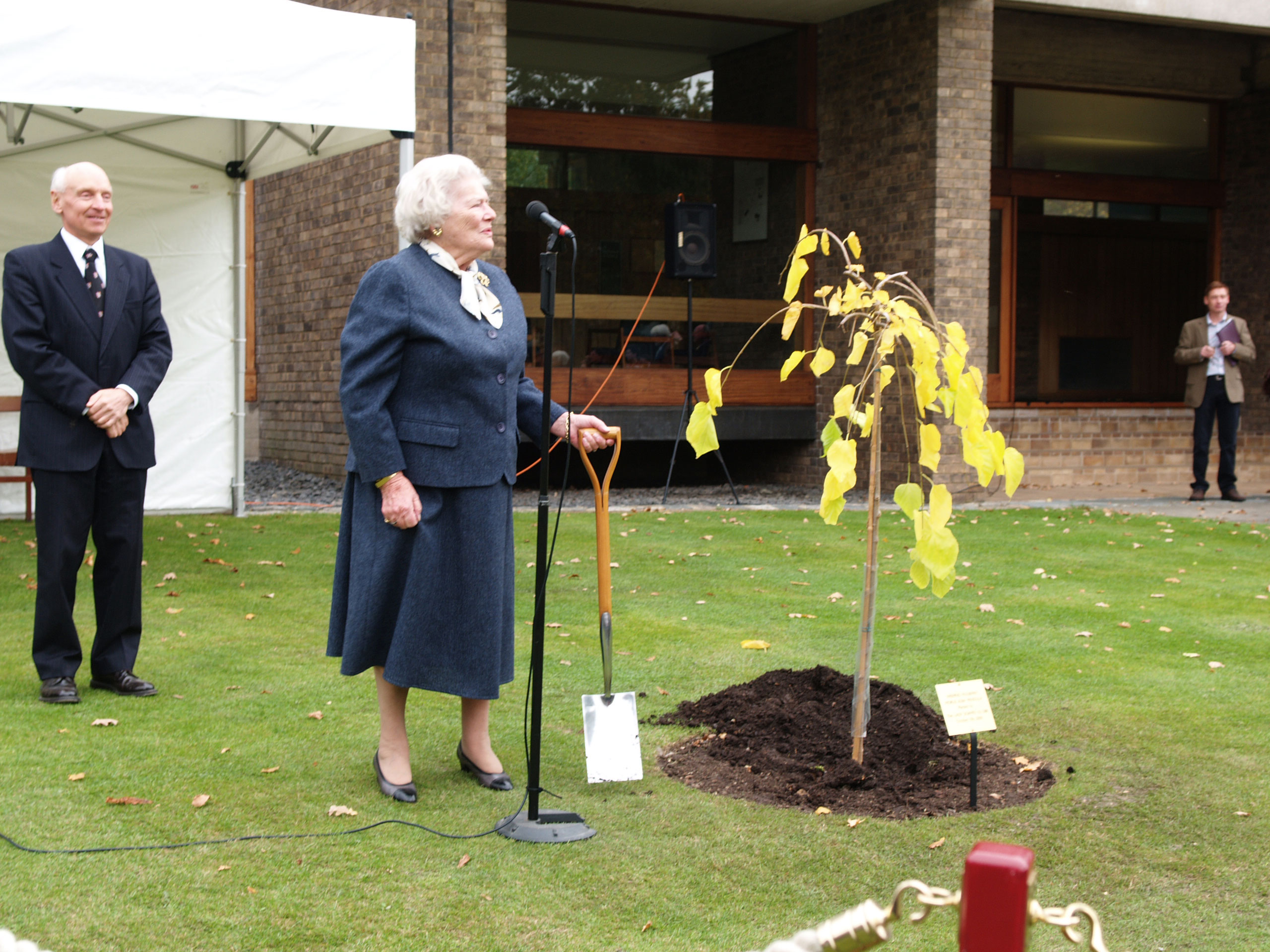 An elderly woman in a dark suit stands at a microphone, holding a shovel next to a newly planted tree. A man in a suit and tie stands nearby. The background includes a brick building and a person observing from a distance.