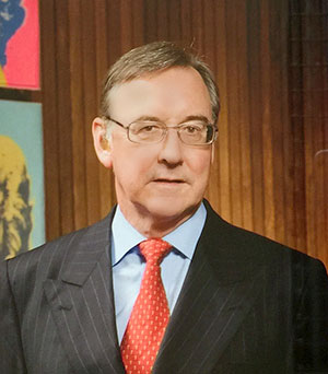 Man in a suit with a red tie and glasses, standing in front of a wooden panel and colorful artwork in the background.