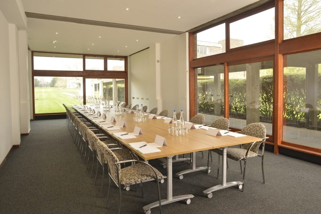 A spacious conference room with a long wooden table set for a meeting. Chairs surround the table, each with a notepad and pen. The room has large windows allowing natural light and offering a view of the greenery outside.