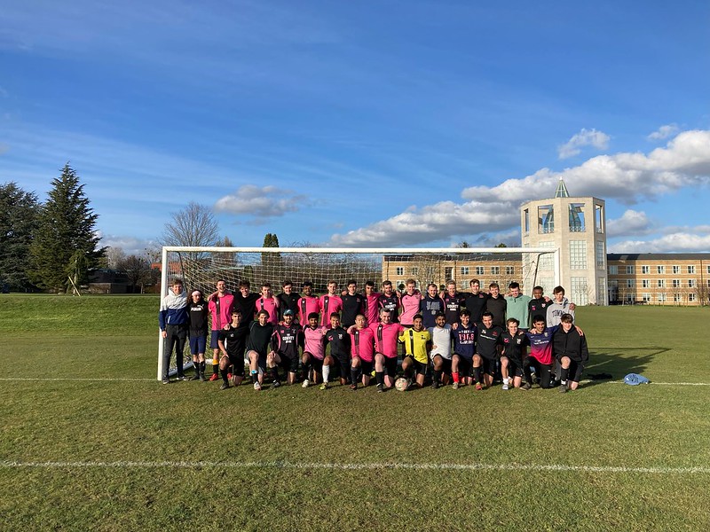 A large group of people, mostly wearing pink and black sports uniforms, pose on a grassy field in front of a soccer goal. A building with a clock tower and trees are in the background under a blue sky with scattered clouds.