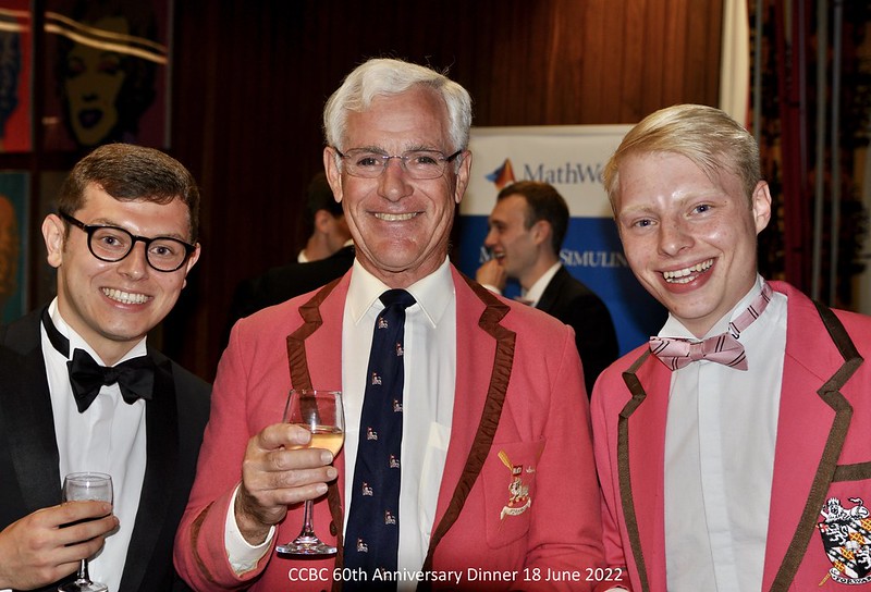 Three people dressed in formal attire, two wearing matching pink blazers, pose for a photo holding drinks. They are smiling and standing indoors at an event. Text on the image reads, CCBC 60th Anniversary Dinner 18 June 2022.