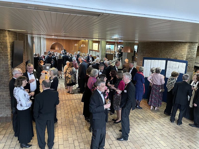 A group of people in formal attire mingle at an indoor event with beige brick flooring. Several individuals are looking at a display board on the right. The setting includes warm lighting and wooden elements.