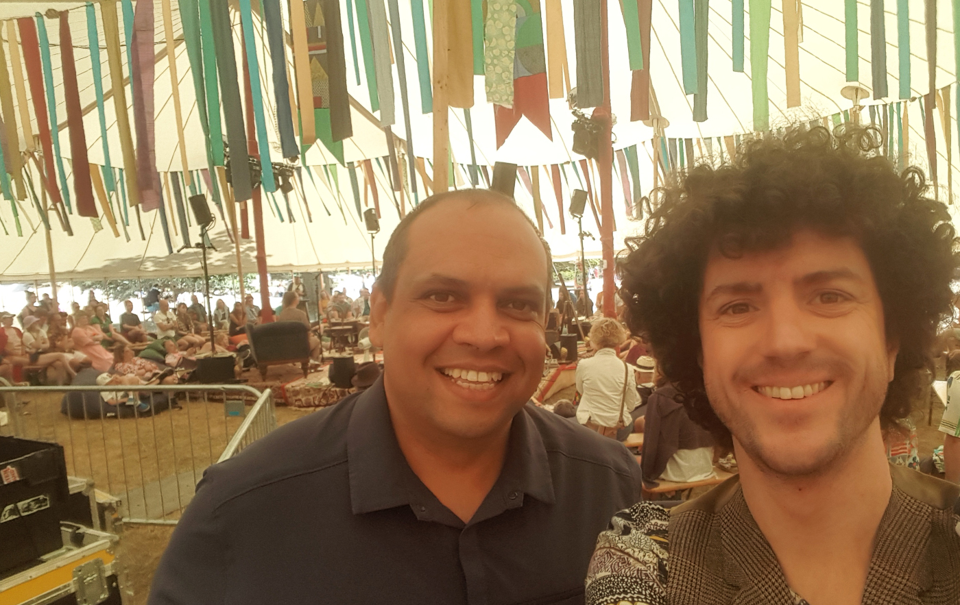 Two men smiling for a selfie in a large tent filled with people. The background is decorated with colorful fabric streamers hanging from the ceiling. The atmosphere appears lively and crowded.
