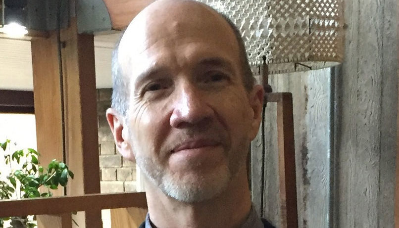 A man with a beard and short hair is smiling slightly. He is wearing a dark jacket and shirt. The background features a wooden wall and a hanging light fixture. A small plant is visible in the corner.