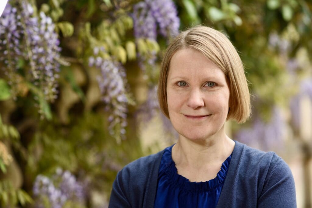 A person with short blond hair and a blue blouse smiles while standing outdoors in front of blooming lavender wisteria vines. The background is softly focused, highlighting the flowers and leafy greenery.