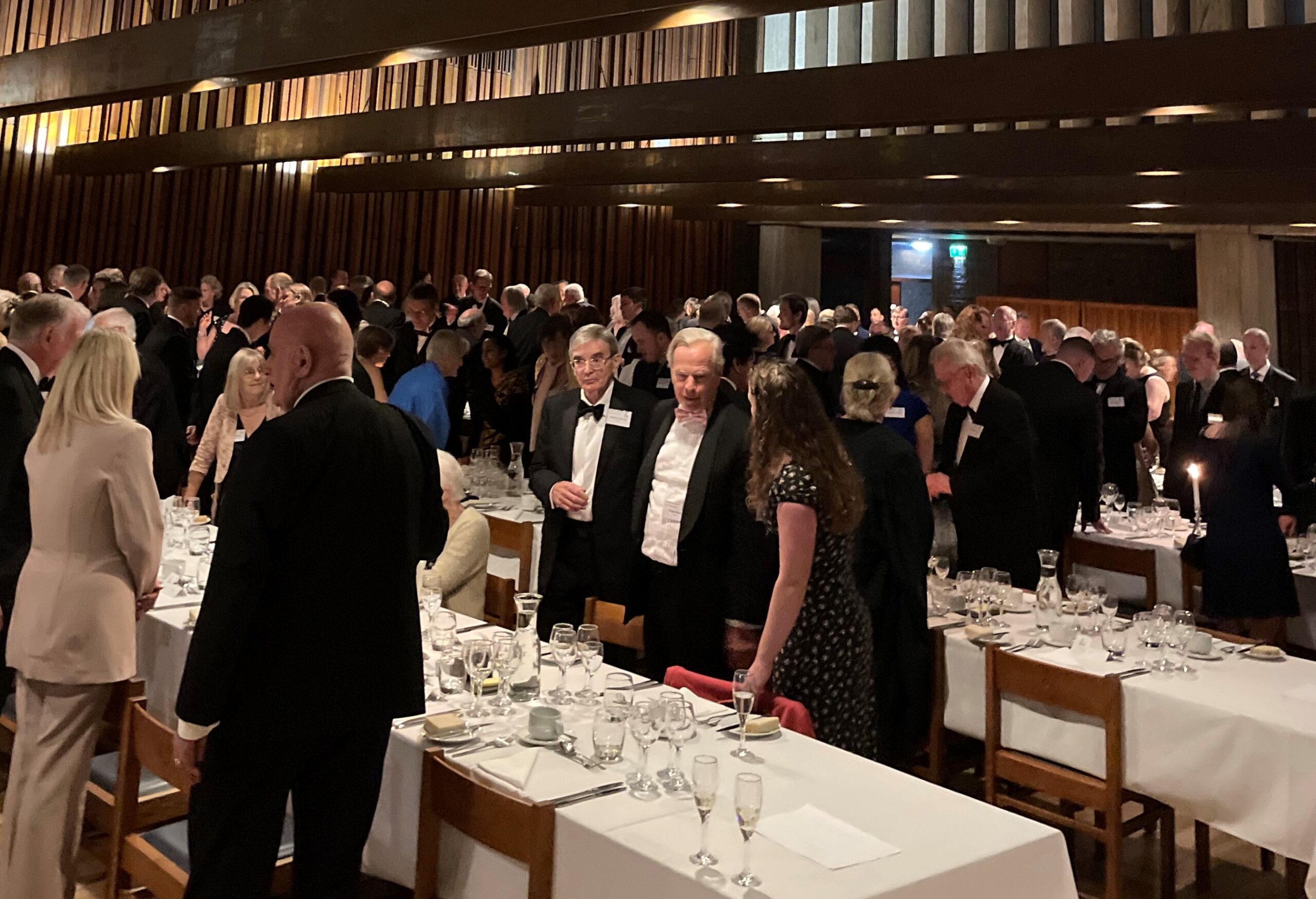 A large group of people in formal attire gather in a banquet hall with wooden decor. Tables are set with white tablecloths, glassware, and cutlery. Candles are lit on some tables, creating a warm ambiance.