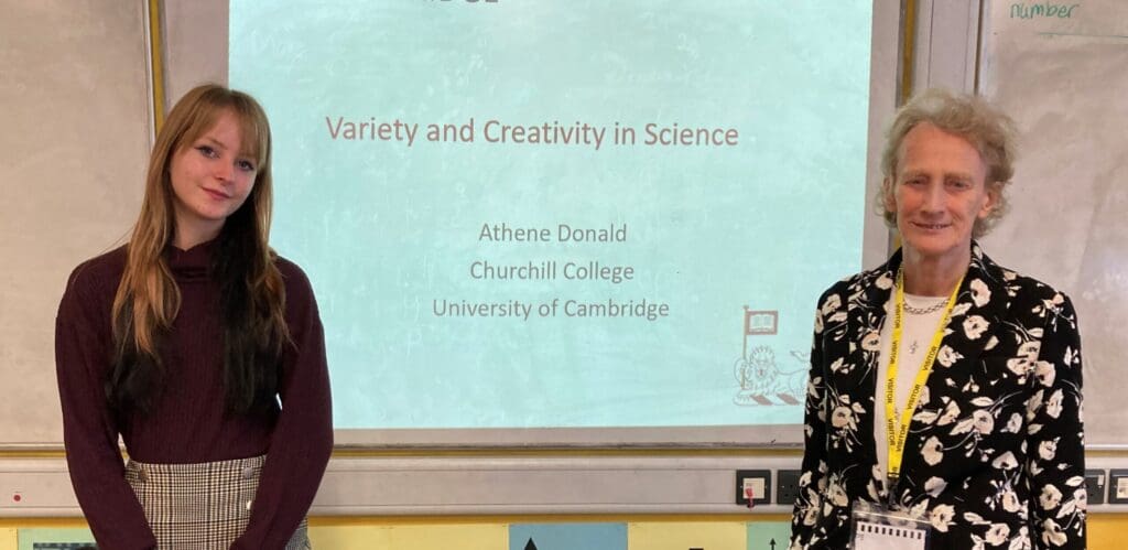 Two people stand in front of a classroom screen displaying a presentation titled Variety and Creativity in Science by Athene Donald at Churchill College, University of Cambridge. The person on the right wears a black and white patterned jacket.