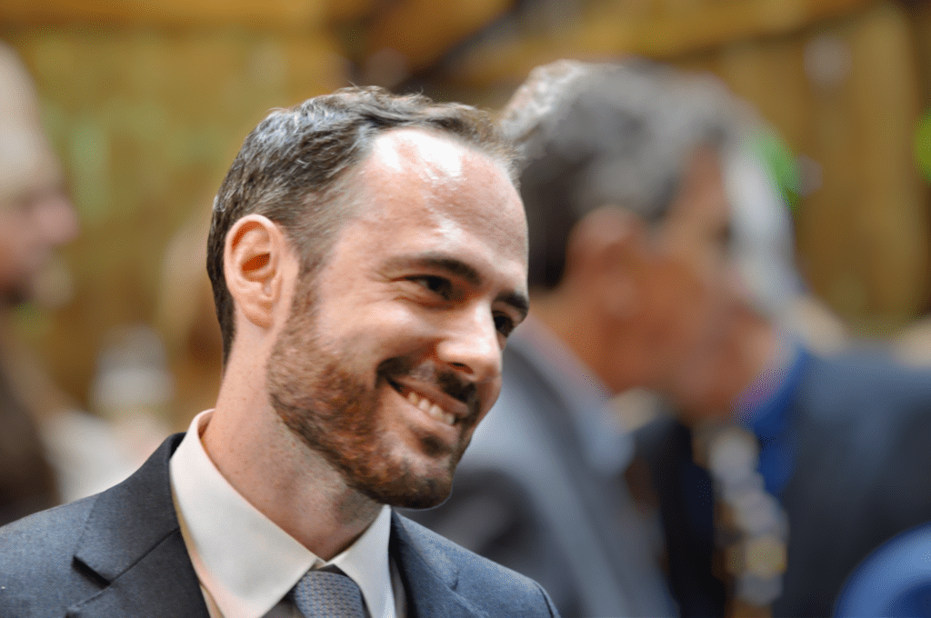 A man in a suit and tie smiles while looking to his left. The background is blurred, with other people in formal attire visible. The setting appears to be an indoor event or gathering.