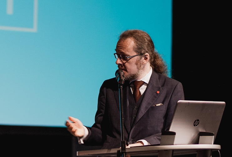 A man with long hair and glasses, wearing a suit and tie, speaks into a microphone at a podium with a laptop. A teal screen is in the background, partially visible.