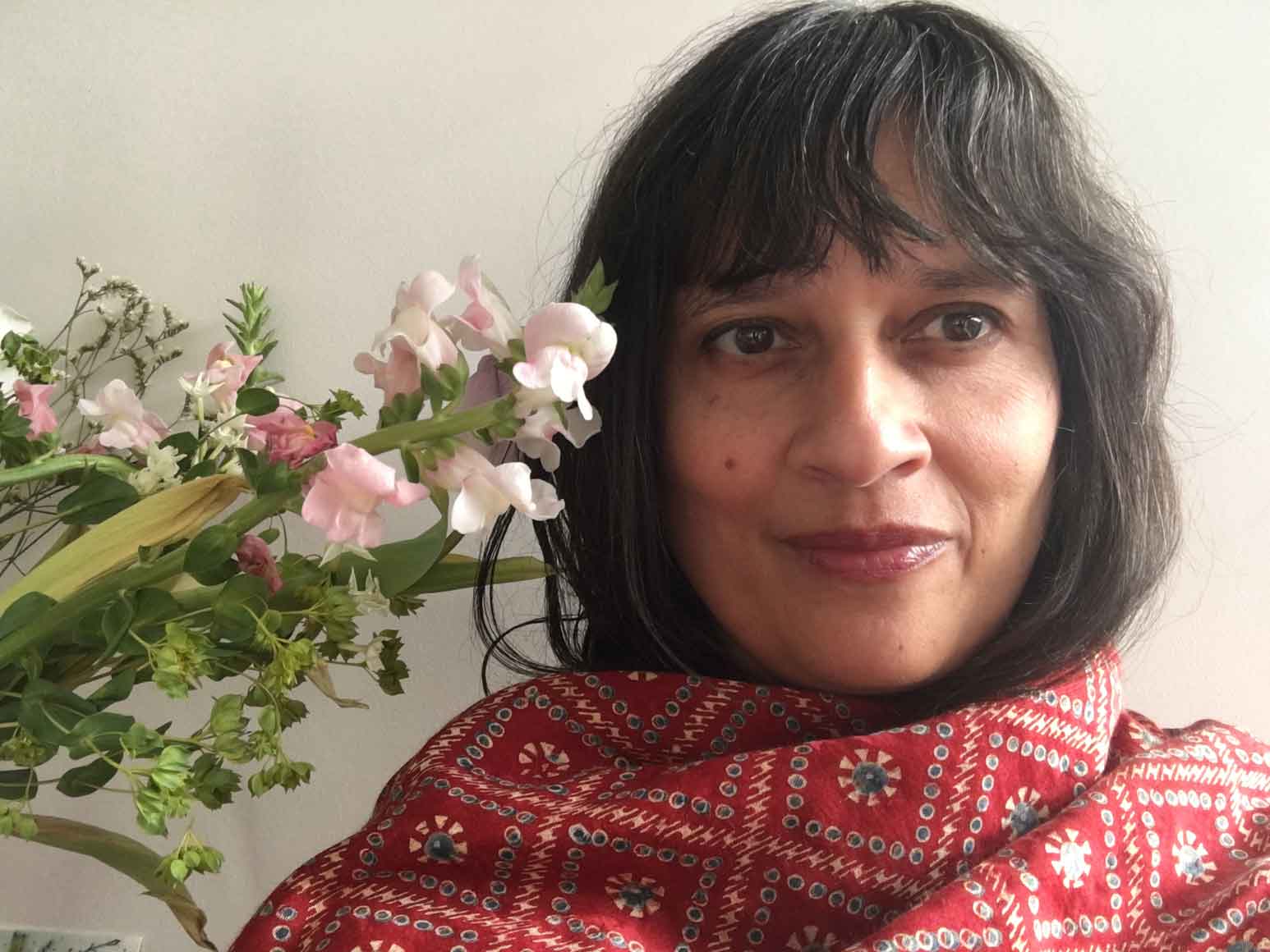 A woman with dark hair and red lip color is wearing a red patterned garment. They are next to a bouquet of flowers with pink and white blooms to their side. The background is plain and light-colored.