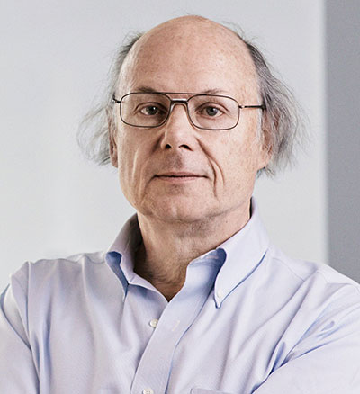 A man with glasses, thinning hair, and a calm expression wears a light blue button-up shirt. The background is blurred and neutral, creating a professional portrait setting.