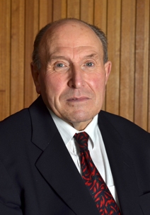 An older man in a dark suit and patterned red tie is sitting against a wooden paneled background, looking directly at the camera.