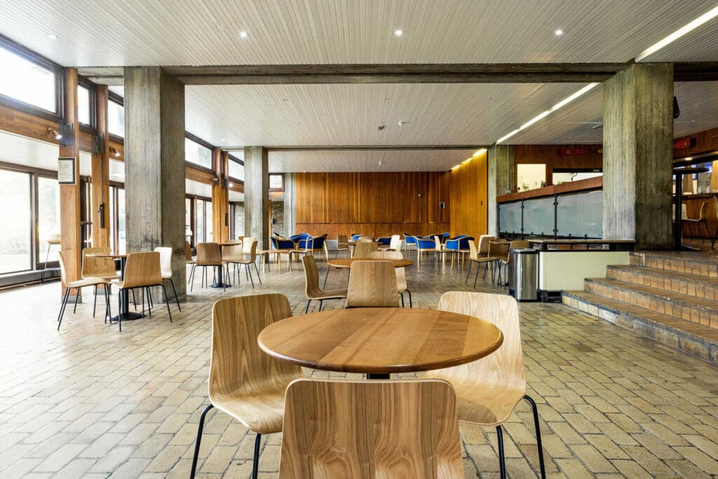 Spacious interior of a dining area with wooden tables and chairs on a tiled floor. Large windows line the walls, allowing natural light to illuminate the room. Modern lighting is installed on the ceiling, with a combination of wood and concrete decor.