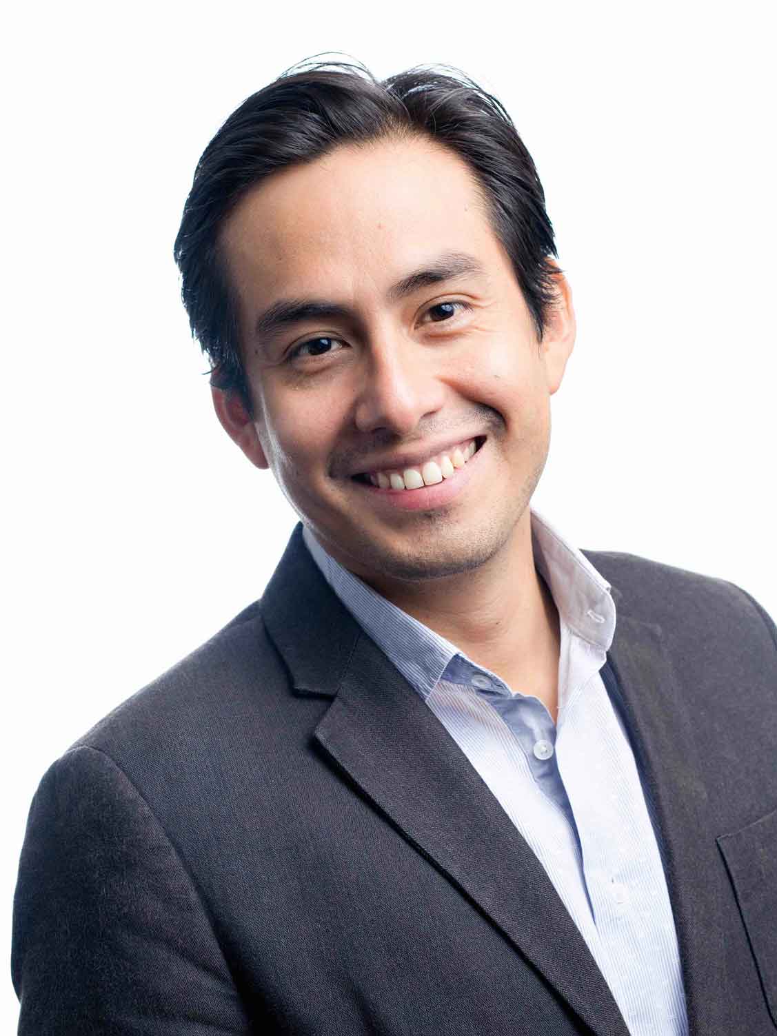 Smiling person with short dark hair wearing a blazer and light-colored shirt, standing against a plain white background.