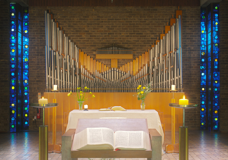 A church altar with an open book and candles is set against a backdrop of grand organ pipes. Blue and green stained glass windows flank the scene. Yellow flowers in vases adorn the altar.