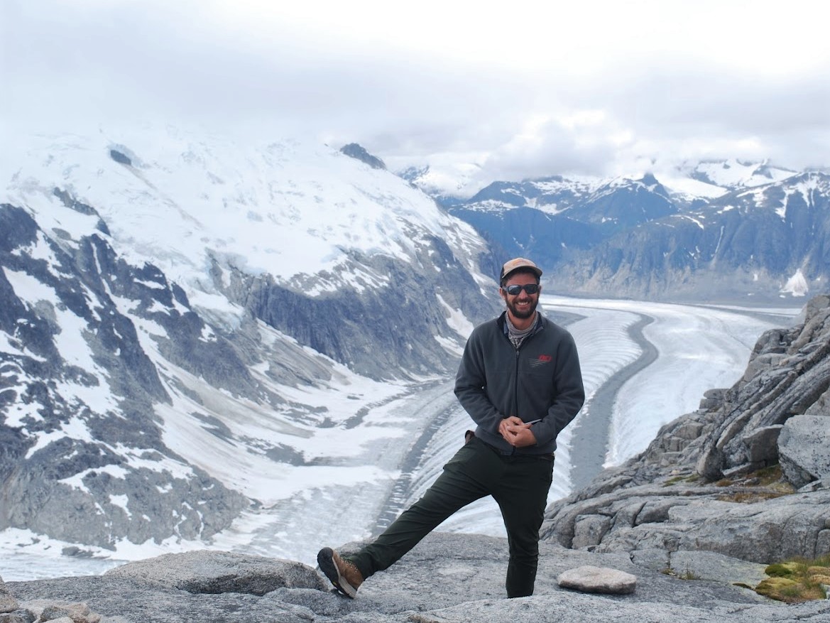 A person wearing sunglasses and a cap stands on rocky terrain, smiling with one leg bent. Behind them, a vast glacier flows between snow-capped mountains under a cloudy sky.