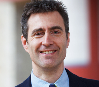 A man with short dark hair is smiling at the camera. He is wearing a dark suit jacket, a light blue shirt, and a light-colored tie. The background is blurred and appears to be an outdoor setting.