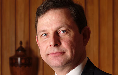 A man in a suit and tie is pictured against a wooden panel background. He has short dark hair and a neutral expression. There is a decorative vase positioned in the background.