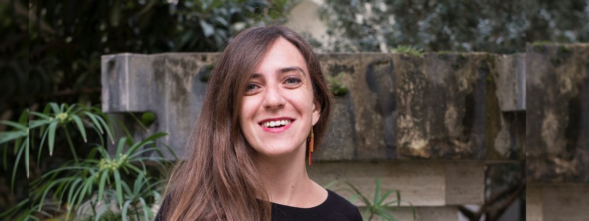 A woman with long brown hair smiling, wearing a black top and orange earrings. She is standing outdoors in front of a stone wall with green plants in the background.