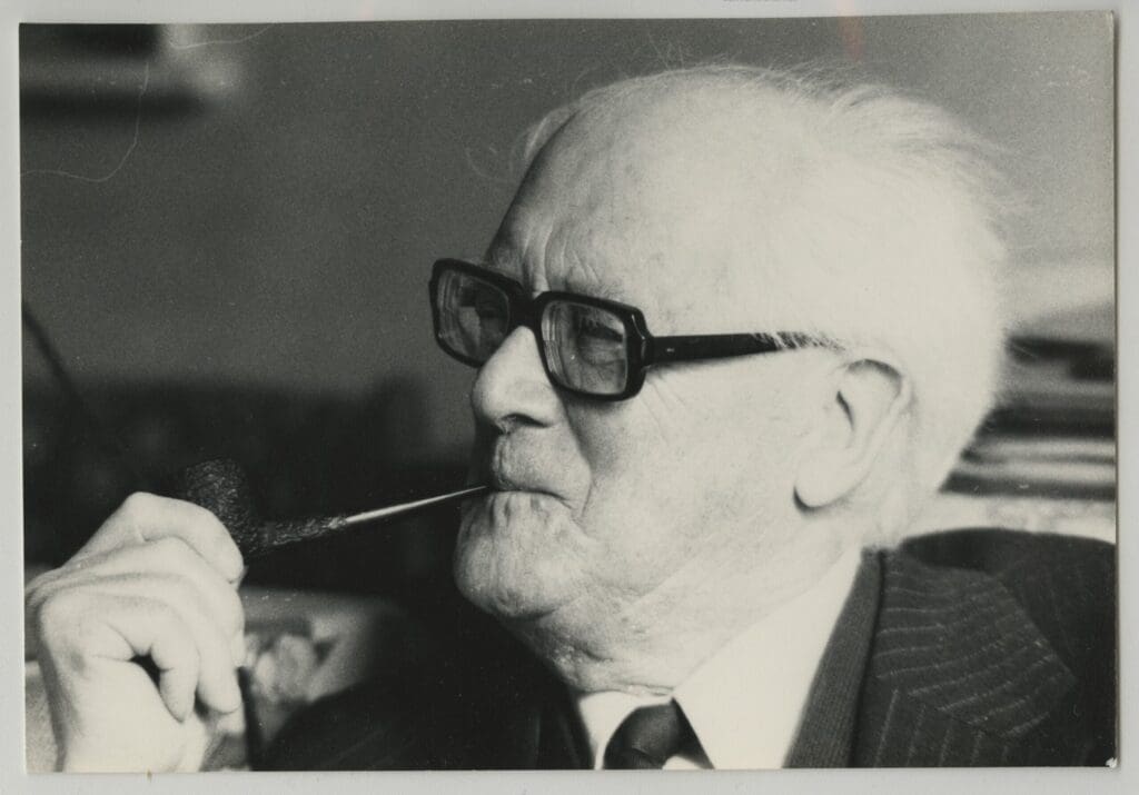 An elderly man with white hair and glasses is sitting and holding a pipe in his mouth. He is wearing a suit and tie, looking contemplatively to the side. The photo is in black and white, suggesting it might be from an earlier era.