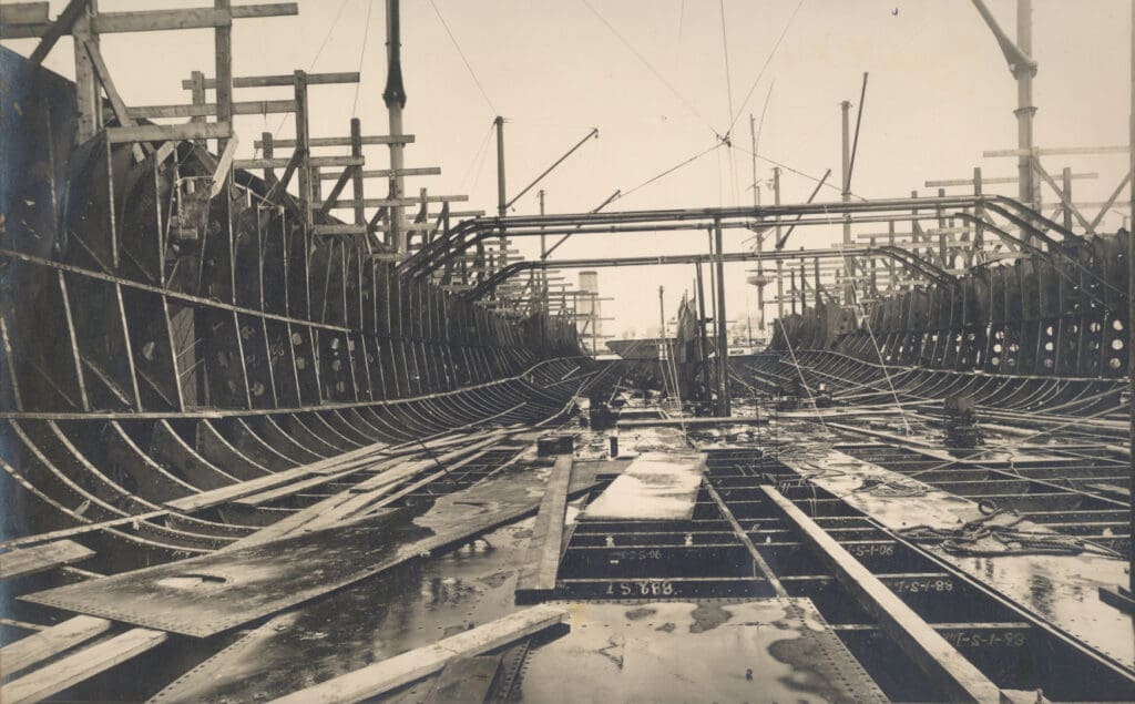 Construction of a large metal ship hull structure in a shipyard. The framework is visible, with horizontal and vertical supports. Scaffolding and beams surround the area, and the ground is wet with some patches of standing water.