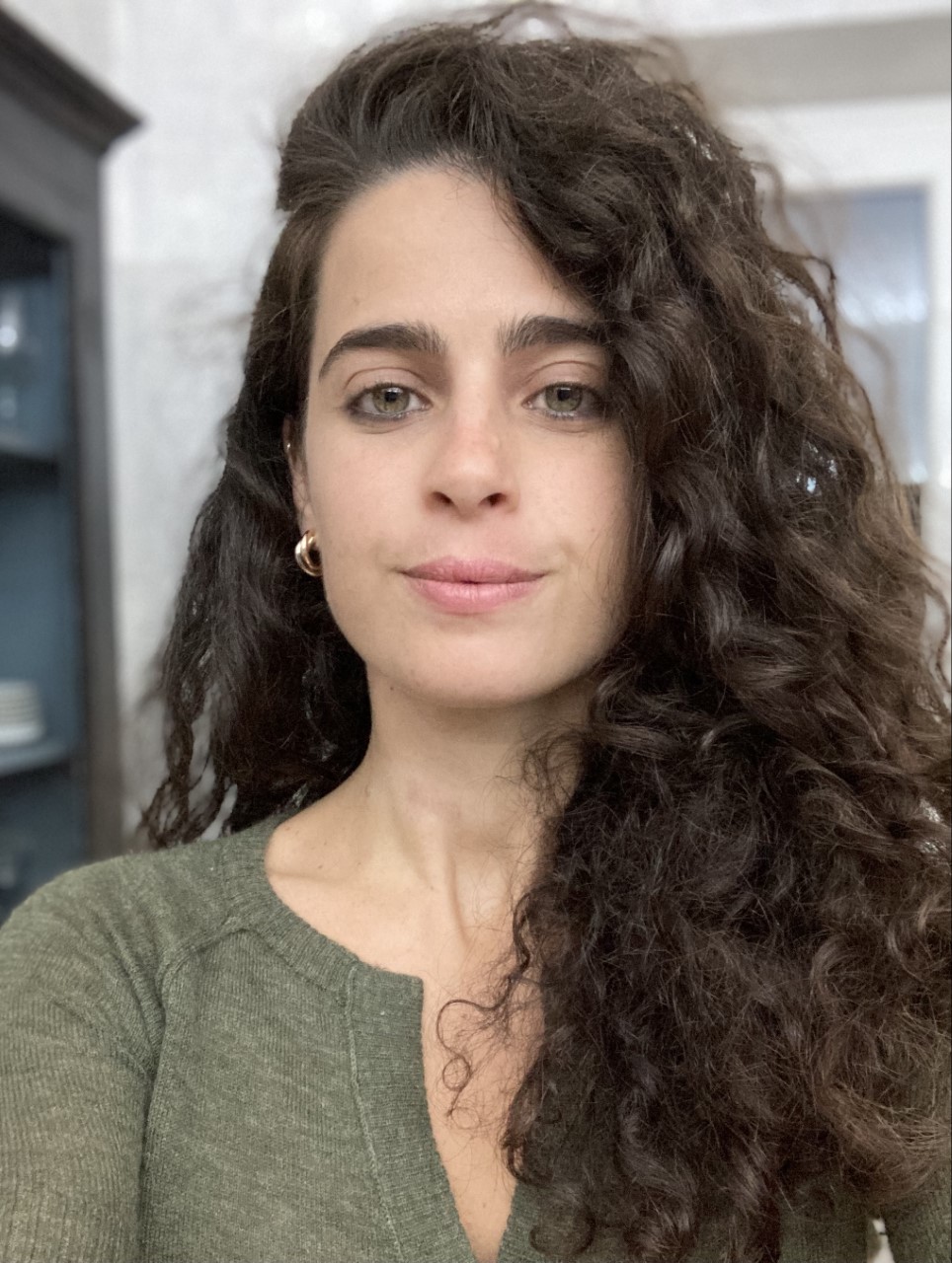 Woman with long, curly dark hair and gold hoop earrings, wearing a green sweater, standing indoors with a neutral expression.