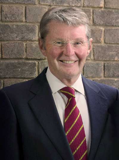 A smiling man with short gray hair wears glasses, a suit, a white shirt, and a striped red and yellow tie. He stands in front of a brick wall.