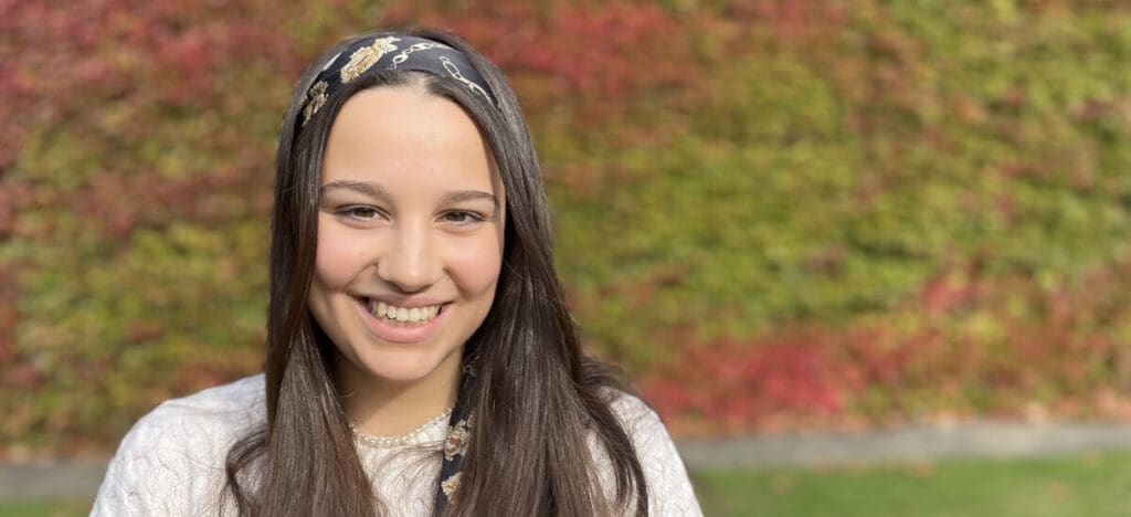 A smiling person with long brown hair, wearing a patterned headband and a light top, stands outdoors. The background is a colorful wall with red and green ivy leaves.