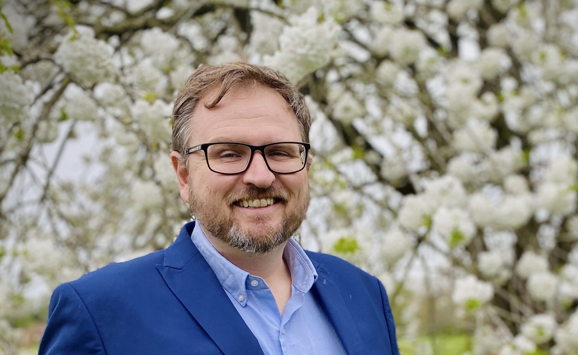 Smiling man with glasses and a beard wearing a blue blazer, standing in front of a tree with white blossoms.