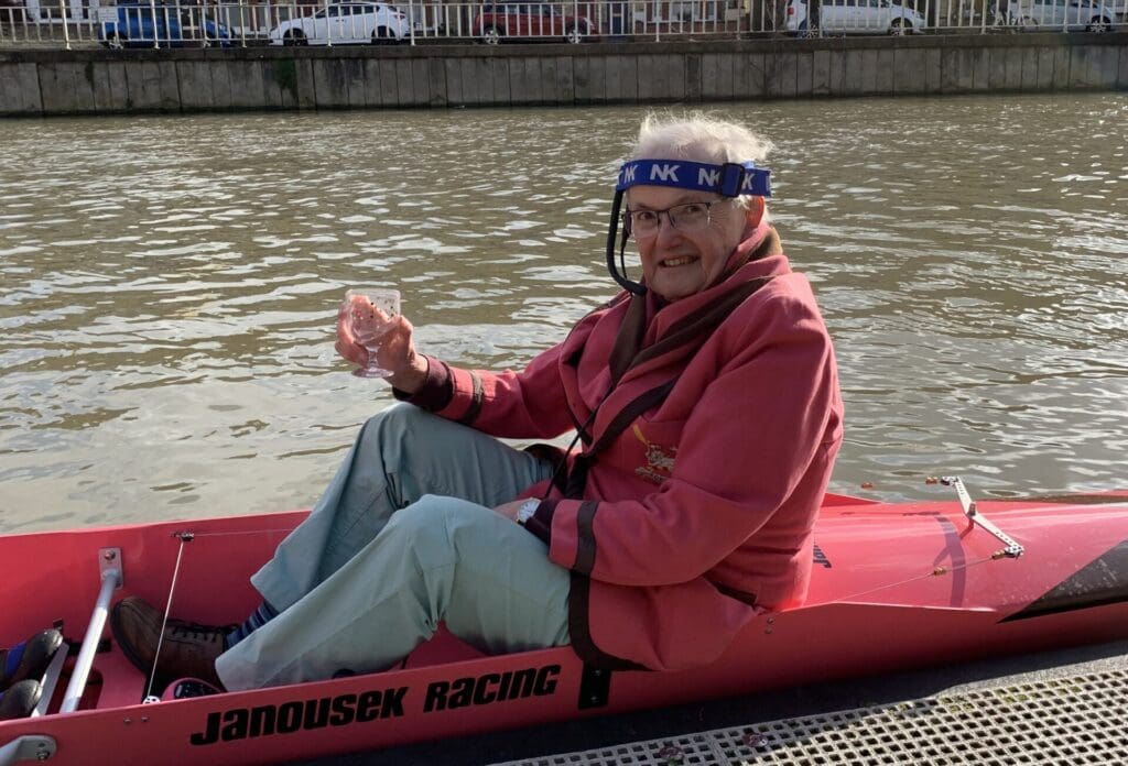 A man sits in a red kayak, holding a glass and smiling. He wears a pink jacket and a blue headband with NK on it. The kayak is stationary on a river near a dock, with buildings and parked cars in the background.