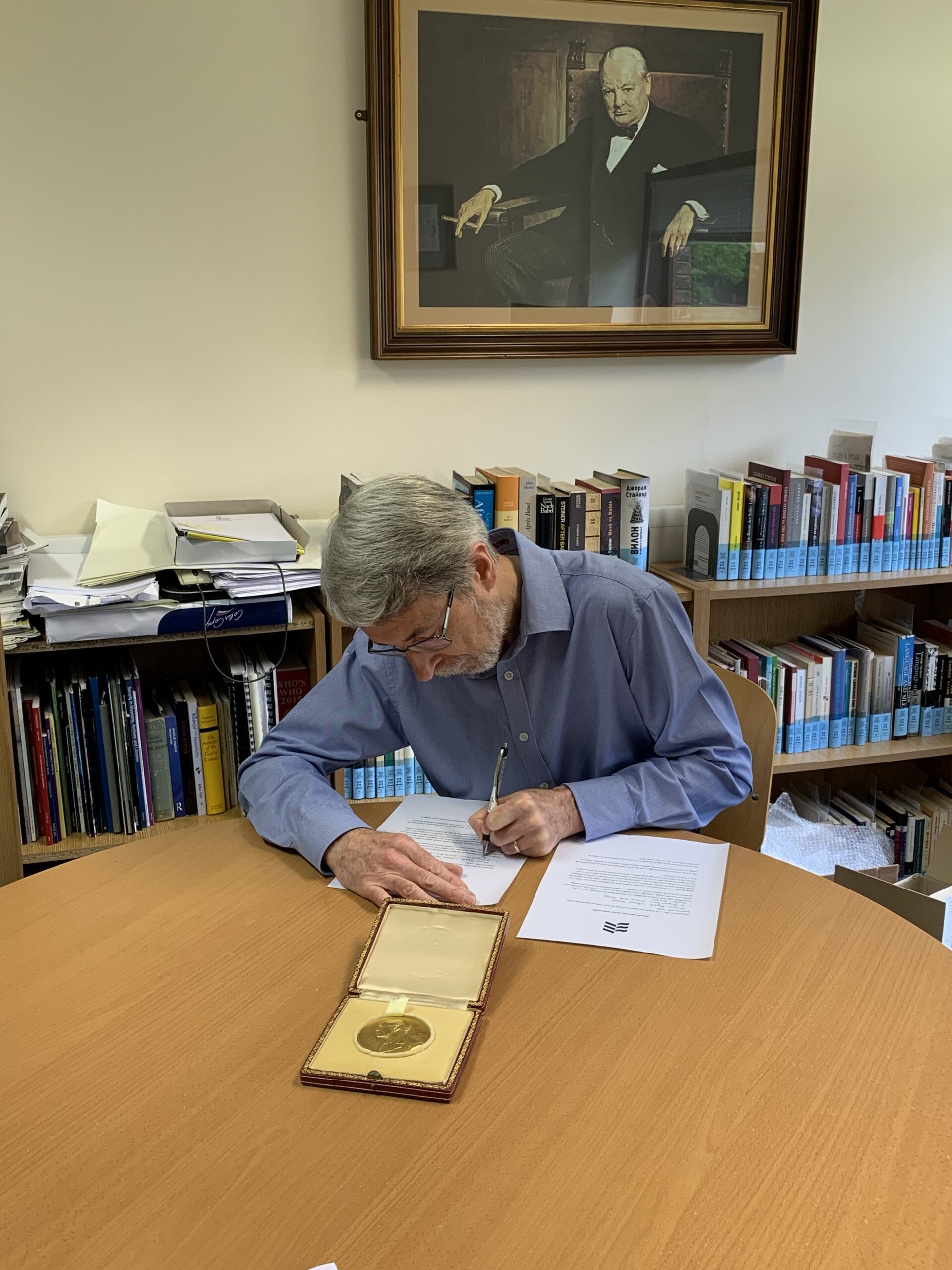 A man with gray hair and glasses is sitting at a table, writing on a piece of paper. A gold medallion in a case is displayed on the table. Books and a painting of another man are in the background.