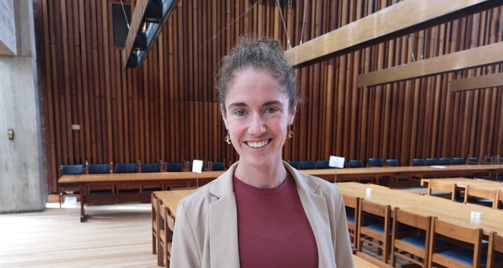 A person with curly hair smiles while standing in a wooden-paneled room with high ceilings. She wears a beige blazer over a maroon top. Long wooden tables and chairs are arranged along the room, and light filters in from above.