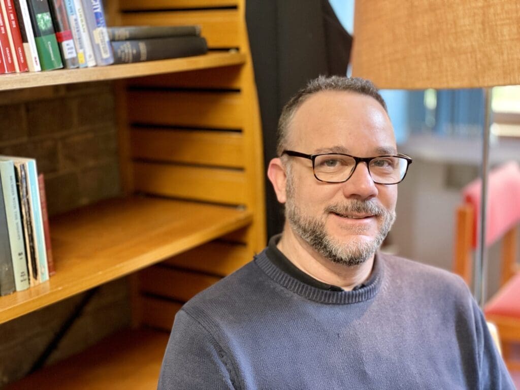 A man with glasses and a beard is sitting in a cozy room. He is smiling and wearing a dark sweater. In the background, there is a bookshelf with books and a warm-colored lamp.