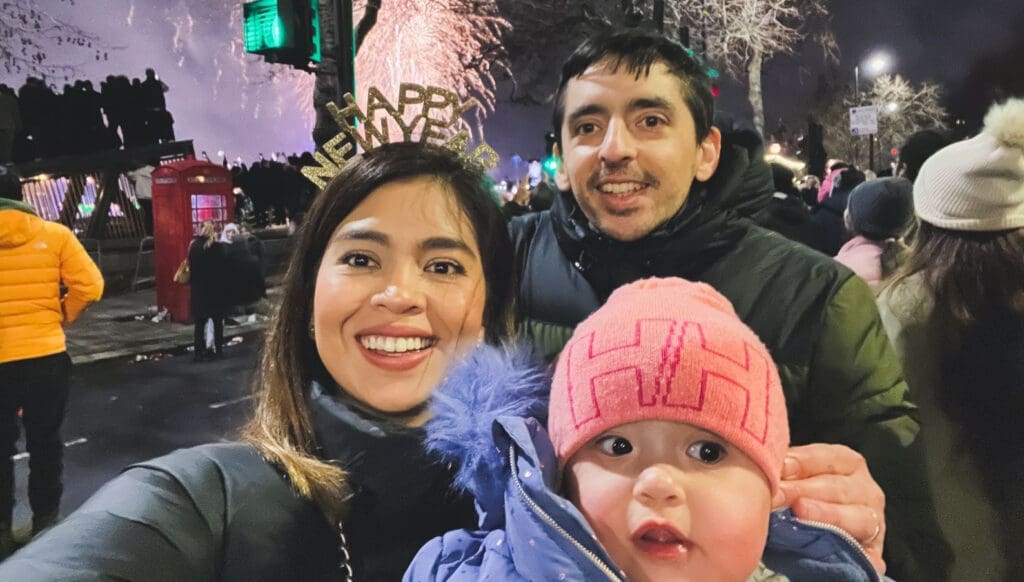 A smiling family at night during New Years celebrations. The woman is wearing Happy New Year headband, and the man is holding a baby in a pink hat. Fireworks light up the sky in the background.