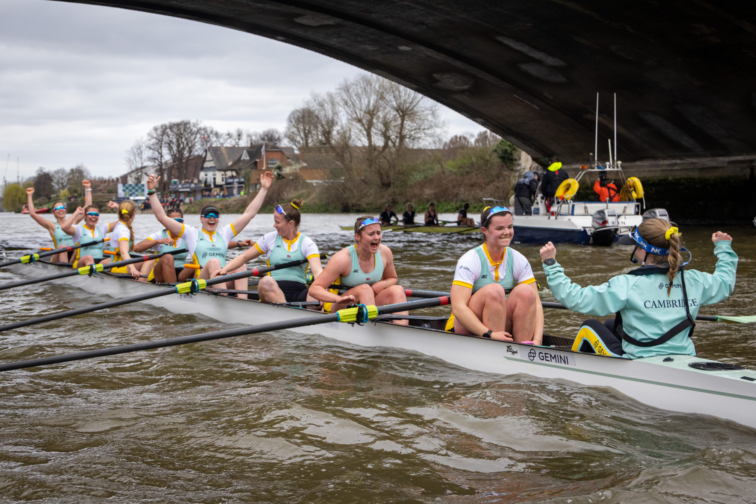 A victorious women's rowing team celebrates as they row under a bridge. They wear yellow and light blue uniforms. The coxswain in light turquoise raises their fist. A support boat follows behind. Trees and houses line the riverbank.