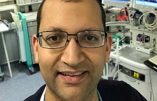 A man with glasses smiling, wearing a dark shirt, in a medical setting with equipment and monitors in the background.