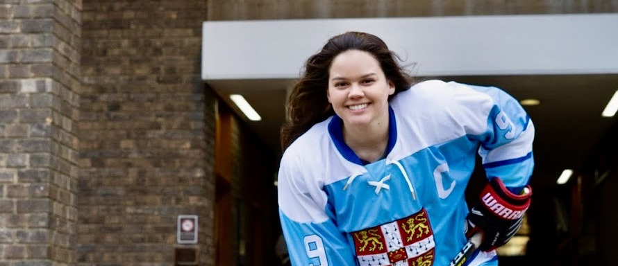 A person in a blue and white hockey jersey, with the number 9 and a C for captain, smiles while holding a hockey stick. She stands in front of a brick building entrance.