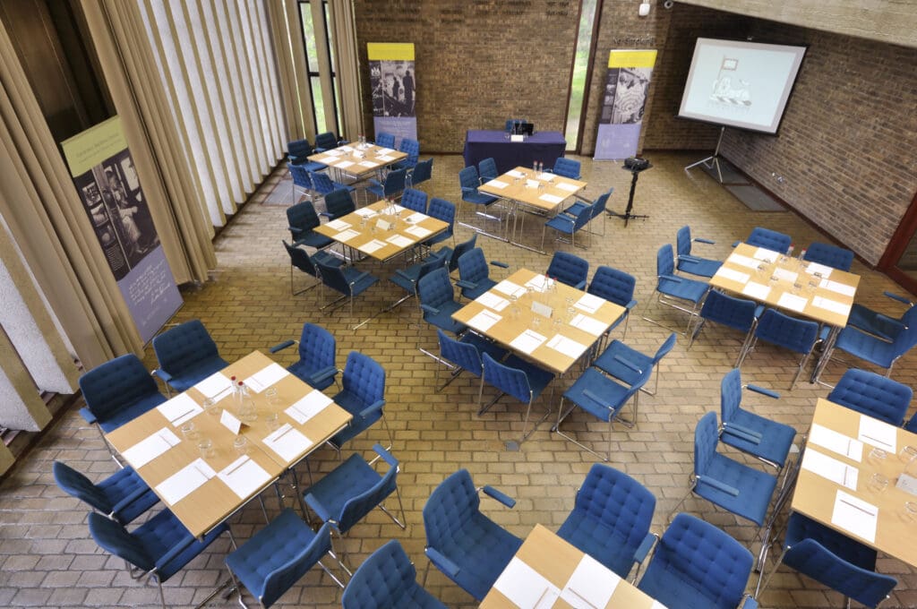 Spacious conference room with brick walls, set up with multiple square tables arranged in clusters. Tables have blue chairs, papers, and glasses, while a projection screen and display banners are at the front. Large windows provide natural light.