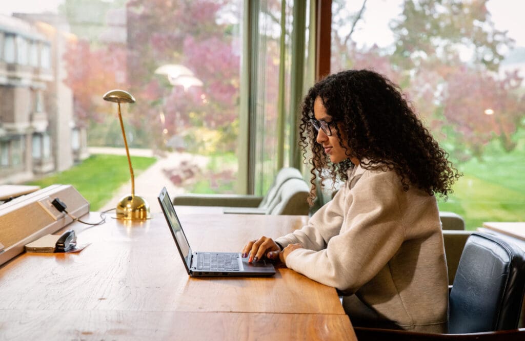 A person with curly hair sits at a wooden desk, wearing glasses and a light sweatshirt, working on a laptop. Behind her is a large window showing a green and purple landscape with trees and buildings.