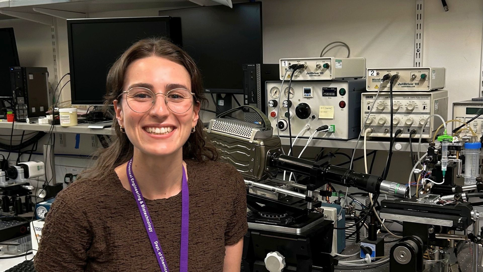 A smiling woman with glasses and long hair stands in a laboratory, wearing a brown top and purple lanyard. Behind her, there is scientific equipment, including monitors, control panels, and various electronic devices.