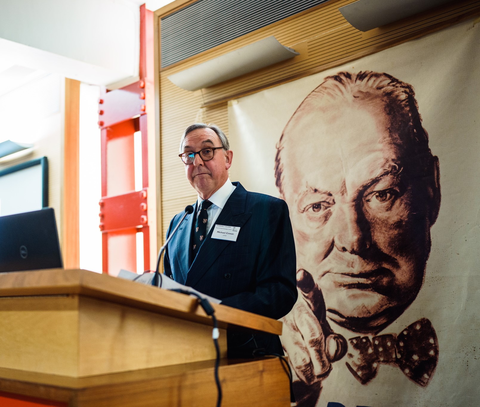 A man in a suit stands at a wooden lectern, speaking into a microphone. Behind him is a large poster of Sir Winston Churchill.