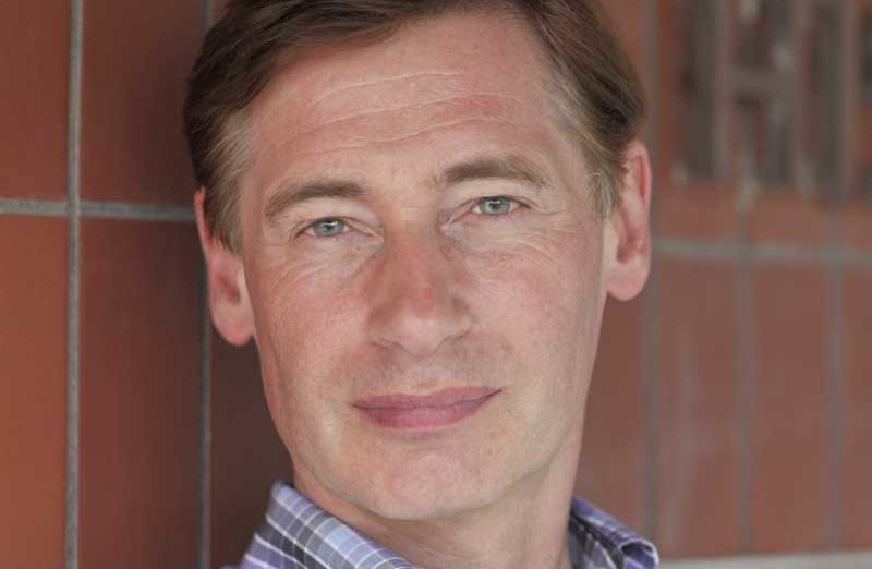 A man with short brown hair is wearing a plaid shirt, standing in front of a tiled wall. They are looking directly at the camera with a neutral expression.