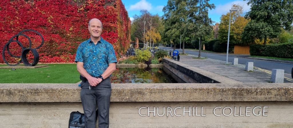 A man stands smiling beside a stone wall with Churchill College engraved on it. Behind him is a pond and a building covered in red ivy. A road with autumn trees lines the right side. A black backpack rests against the wall.