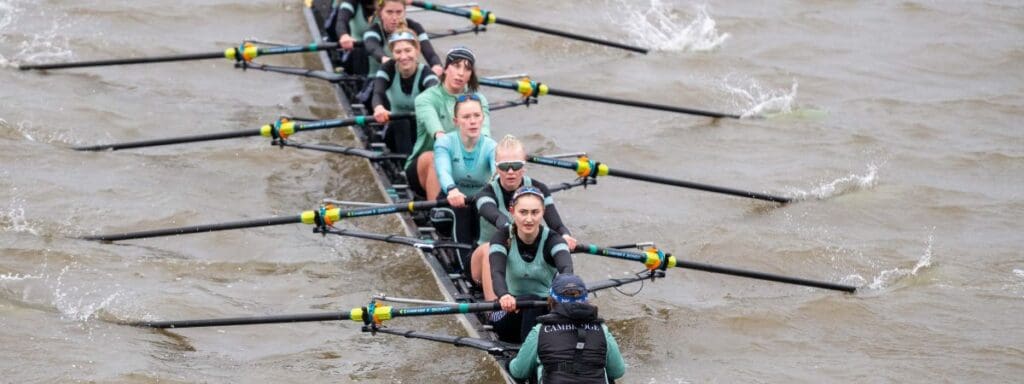 A rowing team of eight, wearing matching teal and black uniforms, is actively rowing on a choppy body of water. The team is coordinated, moving oars in unison, with a coxswain at the front directing them.