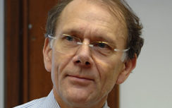 A man with glasses, wearing a striped shirt, looks slightly to the side. The background is blurred, suggesting an indoor setting.