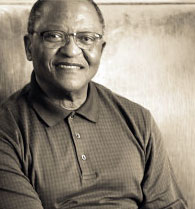 A man is smiling while sitting. He has short hair and is wearing glasses and a collared shirt. The photo is in sepia tones, providing a warm, vintage feel.